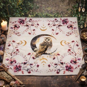 May include: A cream-coloured square tapestry featuring a detailed illustration of an owl perched on a crescent moon. The design includes floral branches with pink flowers and gold crescent moon accents. The tapestry is set on a wooden surface.