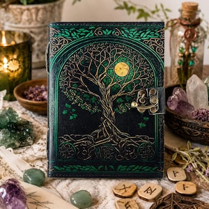 May include: A dark green leather journal with a tree of life design. The cover features a detailed illustration of a tree with a full moon, accented with gold and green. The journal has a metal clasp and is surrounded by crystals and other decorative items.