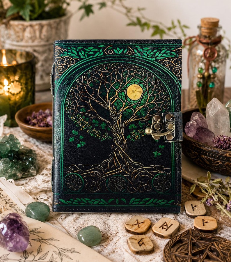 May include: A dark green leather journal with a tree of life design. The cover features a detailed illustration of a tree with a full moon, accented with gold and green. The journal has a metal clasp and is surrounded by crystals and other decorative items.