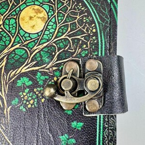 May include: A black leather journal with a tree of life design in green and gold. The cover features a full moon and intricate branches. A metal clasp secures the journal, and a black leather strap is attached.