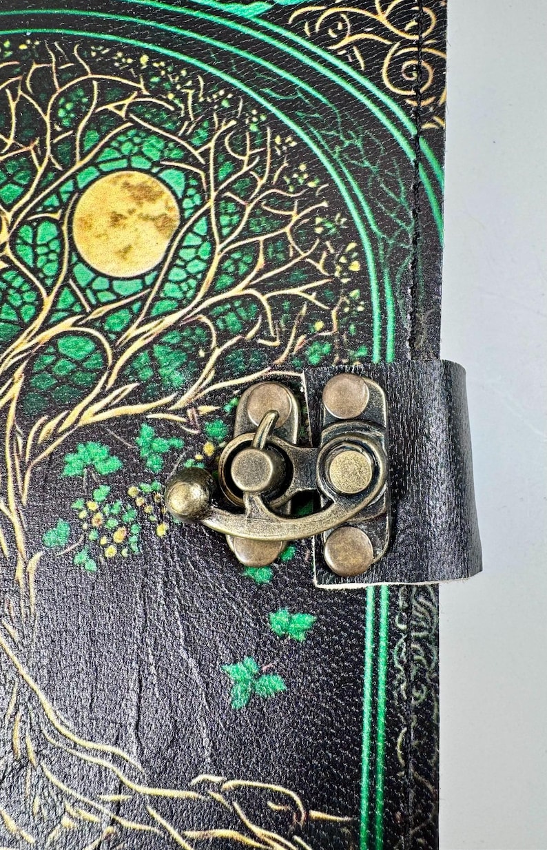 May include: A black leather journal with a tree of life design in green and gold. The cover features a full moon and intricate branches. A metal clasp secures the journal, and a black leather strap is attached.