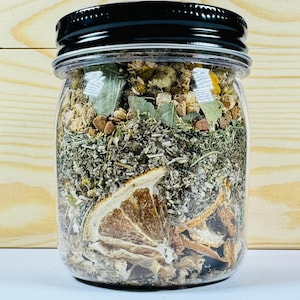 Puede incluir: Un tarro de cristal transparente lleno de hierbas secas, flores y rodajas de naranja, coronado con una tapa negra. El tarro está sobre un fondo de madera clara. El contenido parece ser una mezcla de botánicos, posiblemente para té o un uso similar.