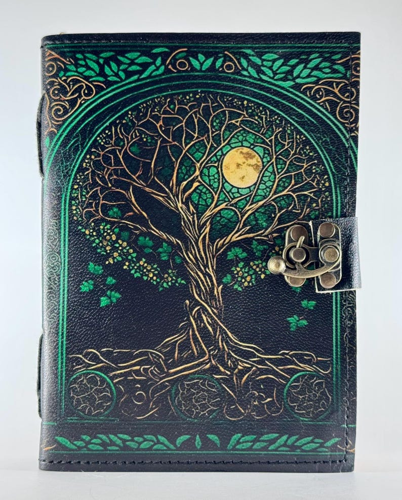 May include: Black leather journal with a tree of life design. The cover features a detailed illustration of a tree with a full moon, in shades of green and gold. It has a metal clasp and a leather strap closure.