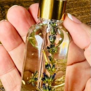 May include: A clear glass dropper bottle filled with a light yellow liquid and a single sprig of dried lavender submerged inside. The bottle features a metallic gold collar and a white rubber dropper top, providing a clear view of the botanical contents.