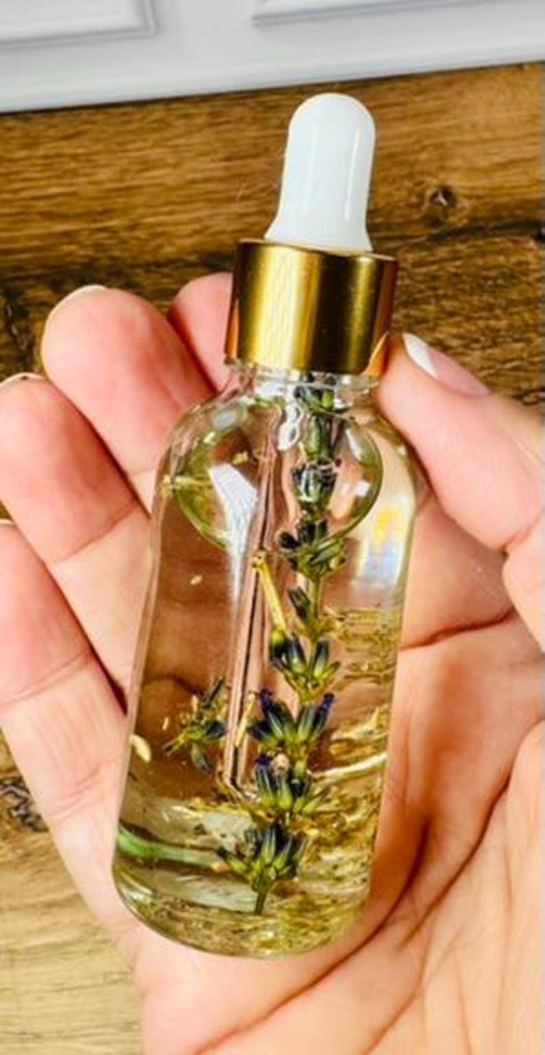 May include: A clear glass dropper bottle filled with a light yellow liquid and a single sprig of dried lavender submerged inside. The bottle features a metallic gold collar and a white rubber dropper top, providing a clear view of the botanical contents.