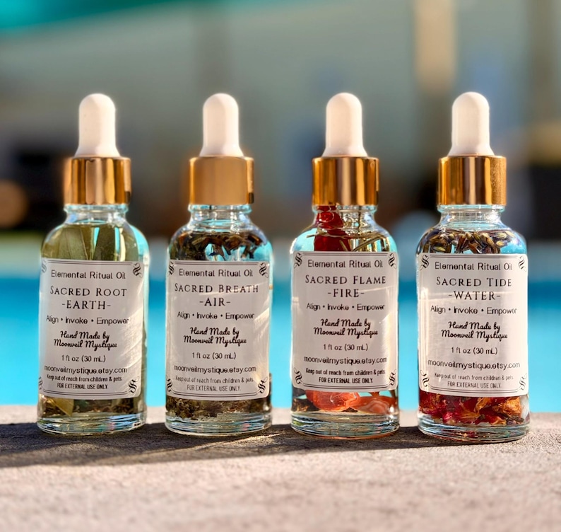 May include: Four clear glass dropper bottles, each containing 30 ml of oil with botanical inclusions. Labels read Elemental Ritual Oil, Sacred Root-Earth, Sacred Breath-Air, Sacred Flame-Fire, and Sacred Tide-Water, with gold-toned dropper caps.