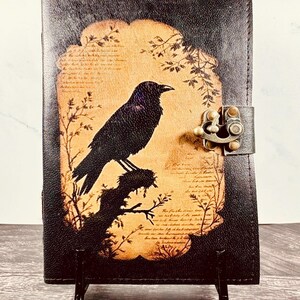 May include: A dark brown leather journal featuring a central tan-colored panel with a black silhouette of a crow perched on a branch. The cover includes faint script text in the background and a metallic bronze-toned latch closure on the right side.