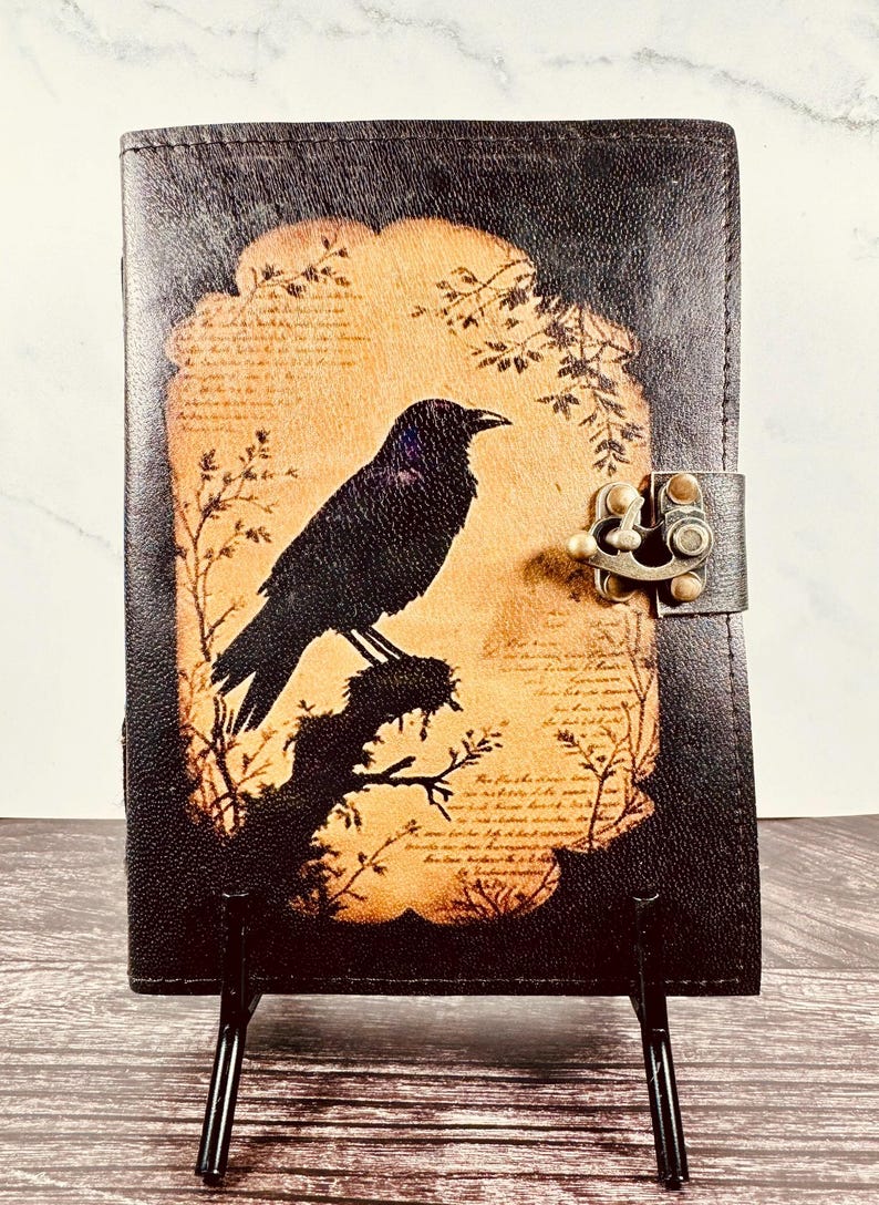 May include: A dark brown leather journal featuring a central tan-colored panel with a black silhouette of a crow perched on a branch. The cover includes faint script text in the background and a metallic bronze-toned latch closure on the right side.