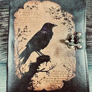 May include: A black leather journal featuring a central tan-colored panel with a printed illustration of a black crow perched on a branch. The design includes faint script text and botanical accents. A metal latch secures the front cover closed.