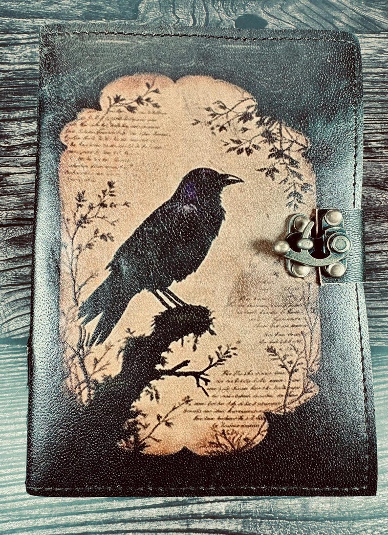 May include: A black leather journal featuring a central tan-colored panel with a printed illustration of a black crow perched on a branch. The design includes faint script text and botanical accents. A metal latch secures the front cover closed.