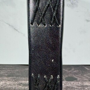 May include: A vertical black leather-like sleeve with a textured surface, featuring two distinct sections of black crisscross stitching. The stitching is arranged in a decorative X-pattern, with small holes punched along the edges of the material.