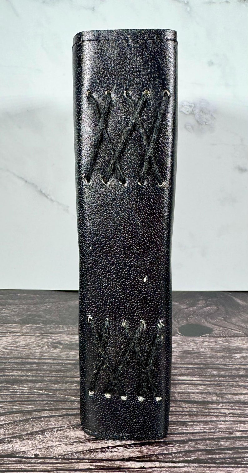 May include: A vertical black leather-like sleeve with a textured surface, featuring two distinct sections of black crisscross stitching. The stitching is arranged in a decorative X-pattern, with small holes punched along the edges of the material.