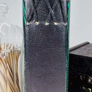 May include: A dark gray leather journal with decorative stitching and a laced closure. The edges are accented with green and gold designs. A glass jar of matches and a small wooden box are in the background.