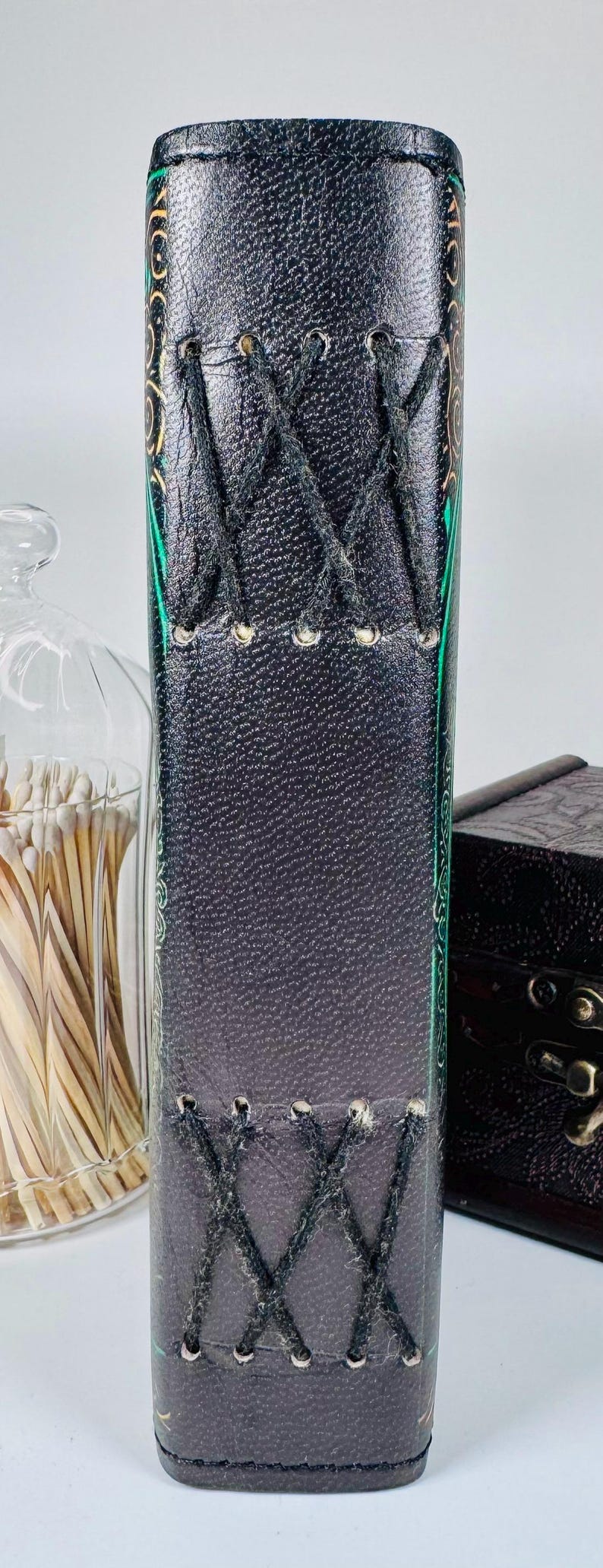 May include: A dark gray leather journal with decorative stitching and a laced closure. The edges are accented with green and gold designs. A glass jar of matches and a small wooden box are in the background.