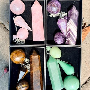 May include: Four black boxes containing polished crystal sets. One box holds rose quartz, one amethyst, one tiger's eye, and one green aventurine. Each set includes a sphere, a pointed tower, a tumbled stone, and a faceted pendulum on a silver chain.