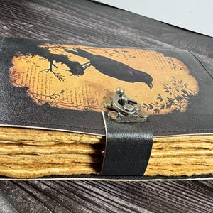 May include: A black leather-bound journal featuring a rectangular graphic of a bird perched on a branch against a tan background with printed text. The journal has thick, textured, light brown deckled-edge pages and a metal latch closure.