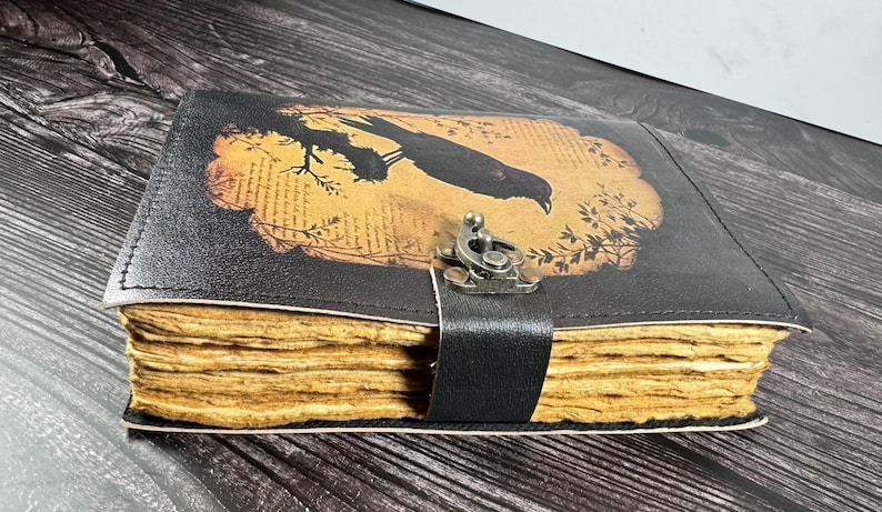 May include: A black leather-bound journal featuring a rectangular graphic of a bird perched on a branch against a tan background with printed text. The journal has thick, textured, light brown deckled-edge pages and a metal latch closure.