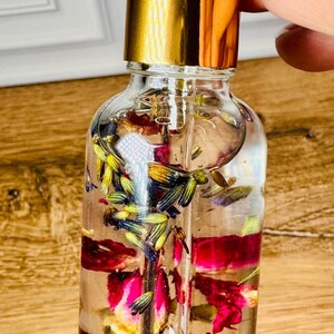May include: A clear glass dropper bottle filled with a transparent liquid containing dried purple lavender buds and vibrant pink rose petals. The bottle features a shiny gold collar and a white rubber dropper top, showcasing the botanical elements inside.