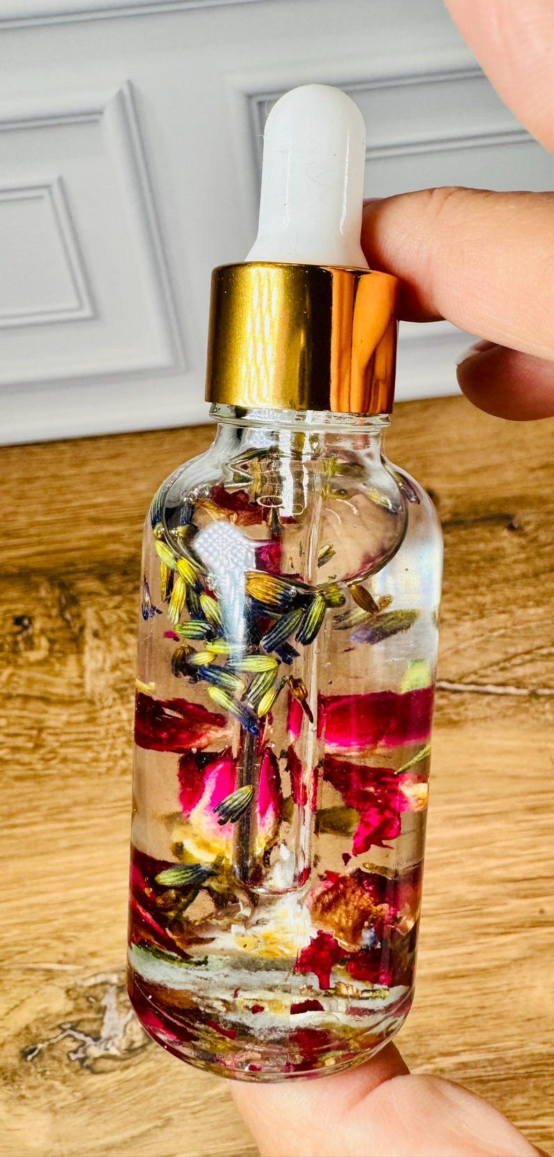 May include: A clear glass dropper bottle filled with a transparent liquid containing dried purple lavender buds and vibrant pink rose petals. The bottle features a shiny gold collar and a white rubber dropper top, showcasing the botanical elements inside.