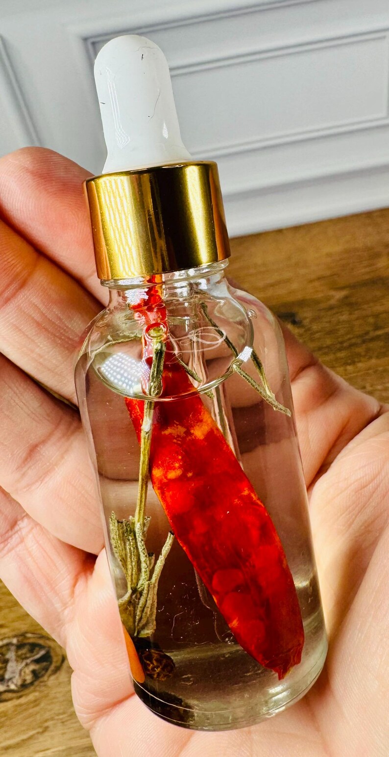 May include: A clear glass dropper bottle with a gold-colored collar and a white rubber bulb. Inside the bottle, a single dried red chili pepper and a small sprig of dried green herb are submerged in a clear liquid, visible through the transparent glass.