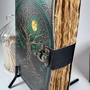 May include: A leather-bound journal with a tree of life design on the cover. The cover is black with green and gold accents. The journal has a black leather strap with a metal clasp. The pages are thick and textured.