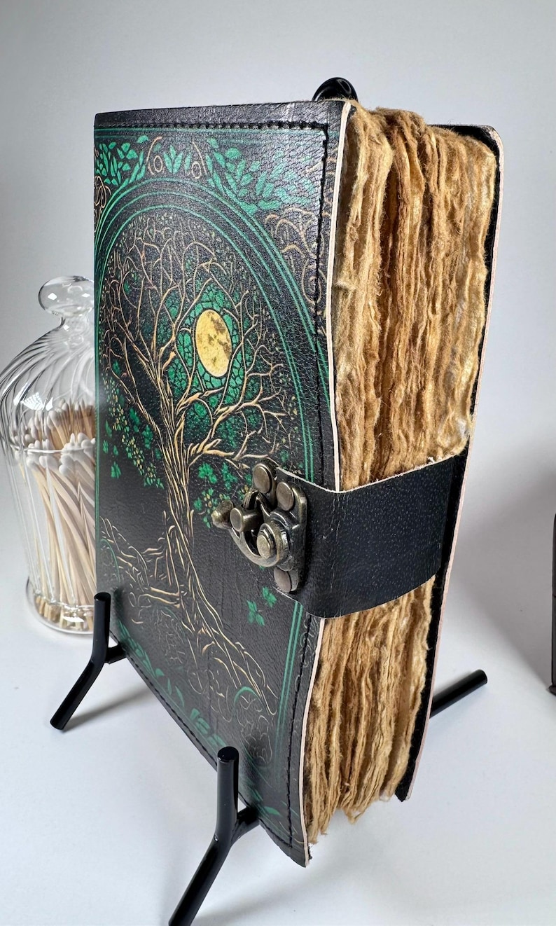 May include: A leather-bound journal with a tree of life design on the cover. The cover is black with green and gold accents. The journal has a black leather strap with a metal clasp. The pages are thick and textured.