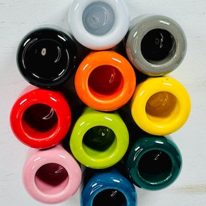 Peut inclure: Une collection de dix objets en céramique en forme d'anneau de différentes couleurs, dont le noir, le rouge, l'orange, le jaune, le vert, le bleu, le rose, le gris et le blanc. Chaque anneau a un centre creux et une finition brillante.