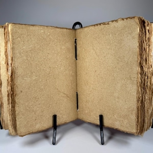 May include: An open, handmade journal with thick, textured, tan-colored pages. The pages are held together with black stitching and bound in a dark cover. The journal is displayed on a black metal stand, showcasing its rustic aesthetic.