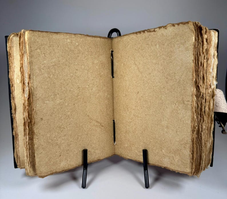 May include: An open, handmade journal with thick, textured, tan-colored pages. The pages are held together with black stitching and bound in a dark cover. The journal is displayed on a black metal stand, showcasing its rustic aesthetic.