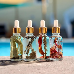 May include: Four clear glass dropper bottles with gold collars and white rubber bulbs. Each bottle contains a different liquid with botanical elements: green leaves, dried herbs, a red chili pepper with rosemary, and colorful dried flower petals.