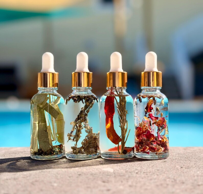 May include: Four clear glass dropper bottles with gold collars and white rubber bulbs. Each bottle contains a different liquid with botanical elements: green leaves, dried herbs, a red chili pepper with rosemary, and colorful dried flower petals.
