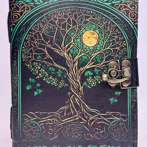 May include: Black leather journal with a tree of life design. The cover features a detailed illustration of a tree with a full moon, in shades of green and gold. The journal has a metal clasp closure.