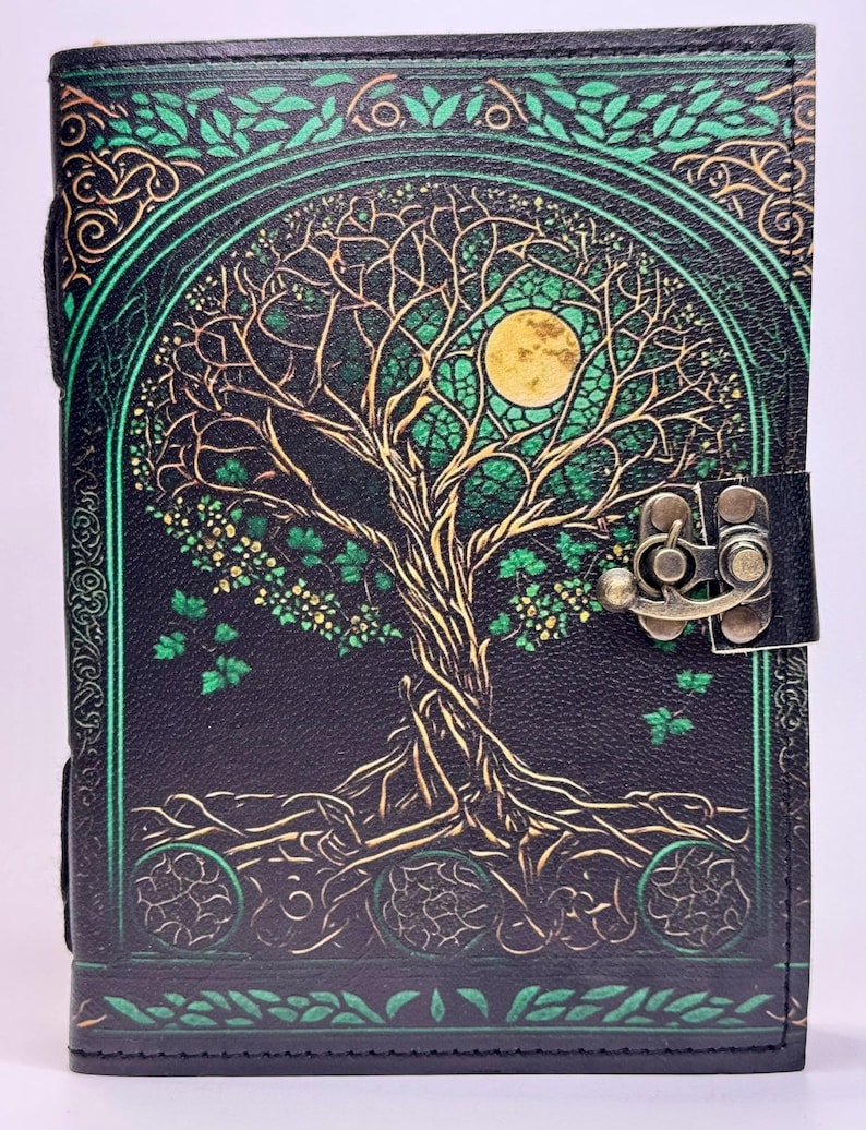 May include: Black leather journal with a tree of life design. The cover features a detailed illustration of a tree with a full moon, in shades of green and gold. The journal has a metal clasp closure.