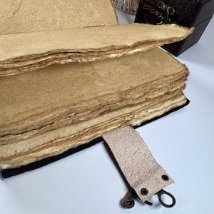 May include: An open, handmade journal with thick, textured, tan-colored pages. The pages have rough, uneven edges. A leather strap with metal hardware is attached to the journal. A small, dark brown wooden box is in the background.