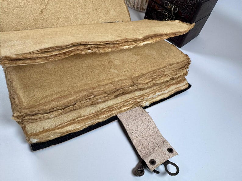 May include: An open, handmade journal with thick, textured, tan-colored pages. The pages have rough, uneven edges. A leather strap with metal hardware is attached to the journal. A small, dark brown wooden box is in the background.