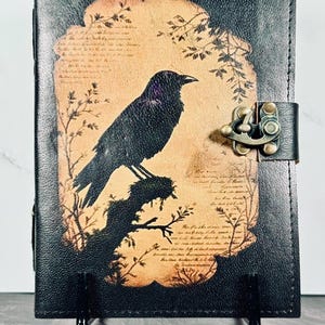 May include: A black leather journal featuring a central tan-colored panel with a printed illustration of a crow perched on a branch, surrounded by faint script text and botanical motifs. The journal is secured with a brass-toned metal latch.