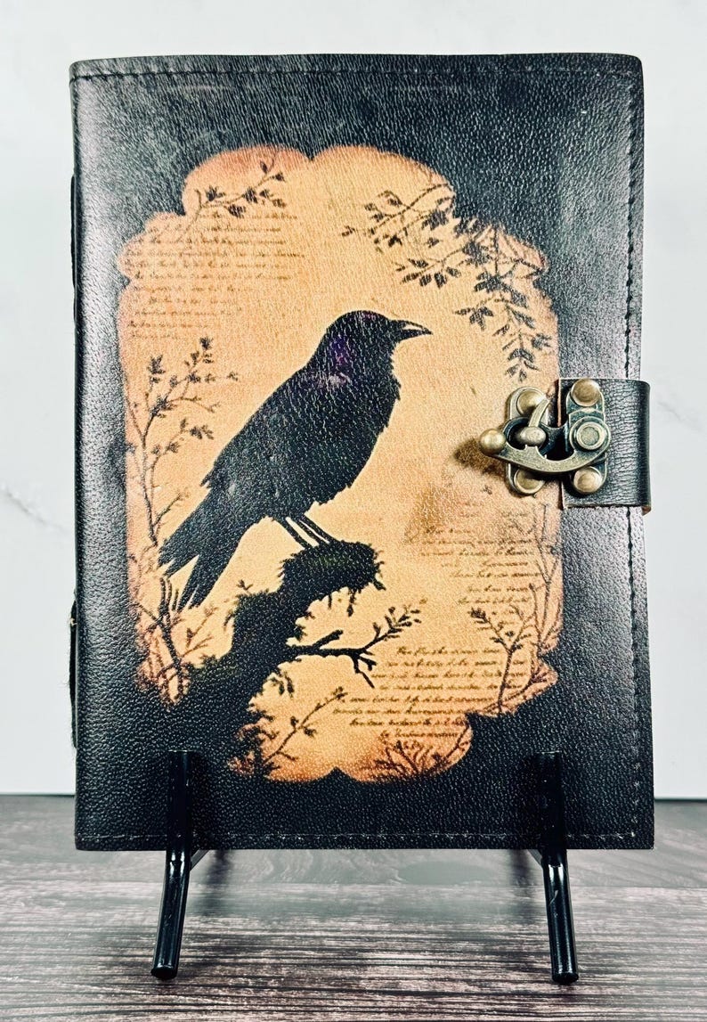 May include: A black leather journal featuring a central tan-colored panel with a printed illustration of a crow perched on a branch, surrounded by faint script text and botanical motifs. The journal is secured with a brass-toned metal latch.