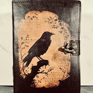 May include: A rectangular black leather journal featuring a central tan-colored panel with a dark silhouette of a crow perched on a branch. The design includes faint script text and botanical illustrations. A metal latch secures the front cover.