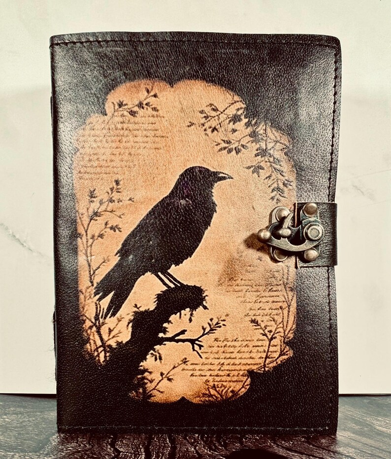 May include: A rectangular black leather journal featuring a central tan-colored panel with a dark silhouette of a crow perched on a branch. The design includes faint script text and botanical illustrations. A metal latch secures the front cover.