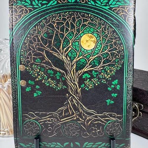 May include: Black journal with a Tree of Life design. The cover features a gold and green illustration of a tree with a full moon. The journal has a black spine and is held by a black stand.