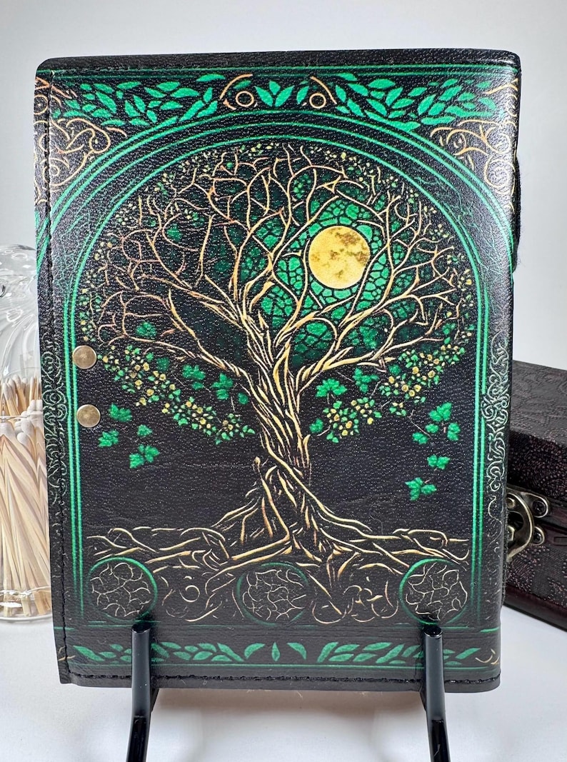 May include: Black journal with a Tree of Life design. The cover features a gold and green illustration of a tree with a full moon. The journal has a black spine and is held by a black stand.