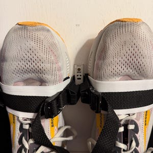 May include: Pair of athletic shoes with white mesh uppers, pink and yellow accents, and white laces. The shoes are secured with black straps and buckles. The brand name "BROOKS" is visible on the side of the shoes.