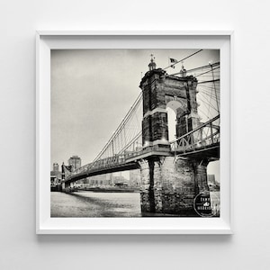 May include: Monochrome photograph of a suspension bridge featuring a stone tower and archway. The bridge stretches across water, with a city skyline in the background. The image is framed within a white square.