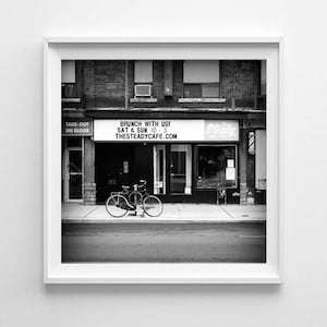 May include: Monochrome image of a cafe exterior with a bicycle parked outside. A sign above the entrance advertises "BRUNCH WITH US! SAT & SUN 10-3 THESTEADYCAFE.COM". The building is brick with a "TAKE-OUT ON BLOOR" sign.