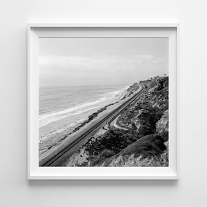 May include: Monochrome photograph of a coastal scene. A railway line follows the coastline, next to a sandy beach and the sea. The image is framed in a white square frame.