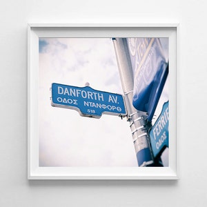 May include: A framed photograph featuring a blue street sign with the text "Danforth Ave" in English and Greek. The sign is fixed to a metal pole, set against a cloudy sky. The number "518" is also visible on the sign.
