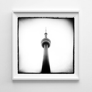 May include: Monochrome photograph of the CN Tower in Toronto, Canada, presented within a white frame. The tower's unique structure is centrally positioned against a clear sky. The image has a vintage, slightly grainy appearance.
