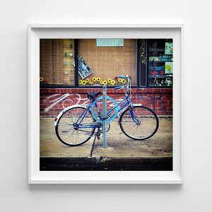 May include: Framed photograph of a blue bicycle parked on a pavement. The bicycle is secured to a metal post with a blue circular lock. The background includes a brick wall, a shop window, and graffiti.
