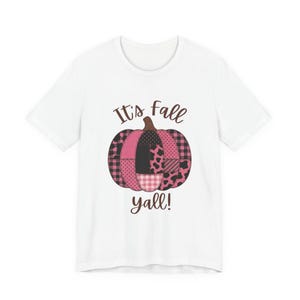 Peut inclure: Un t-shirt blanc à manches courtes avec un motif central représentant une citrouille composée de divers empiècements roses et noirs, incluant des carreaux, des pois et des motifs animaliers, avec le texte marron It's fall yall! inscrit.