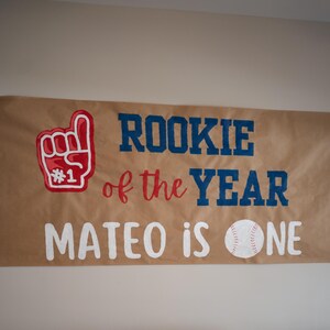 May include: A baseball-themed party setup with a banner that reads "Rookie of the Year" and "Mateo is One." The banner features a foam finger and baseball graphics. A table is set with food and a baseball-themed tablecloth.